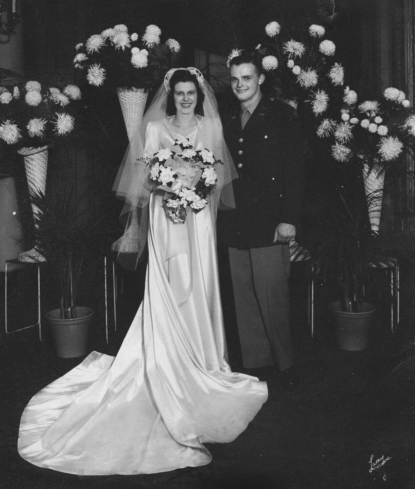 Black-and-white wedding portrait of Jack in uniform beside Jean in her gown and veil, surrounded by floral arrangements, on their wedding day in January 1944.