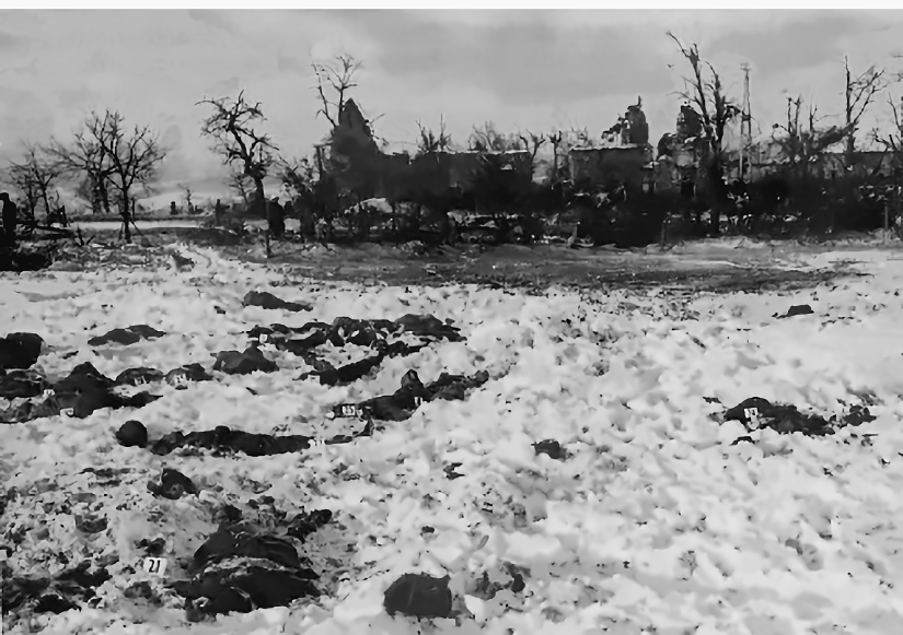 Snow-covered field near Malmedy after the massacre, with the bodies of American soldiers in the snow and damaged buildings behind.