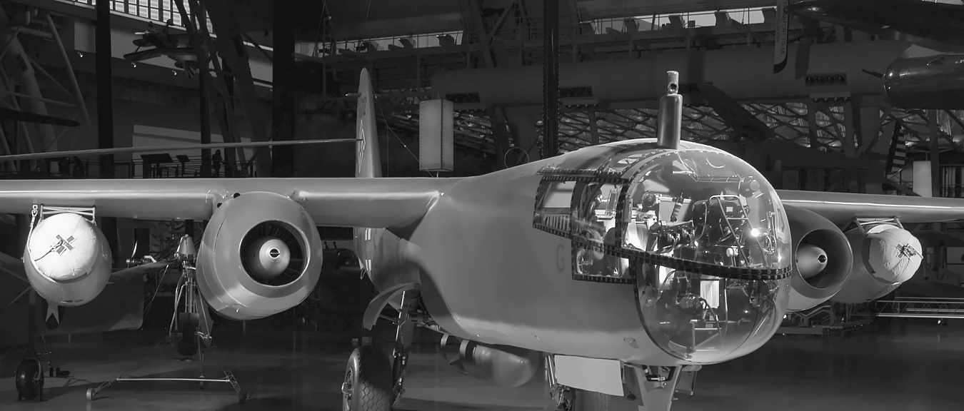 Black-and-white photograph of an Arado Ar 234 twin-engine jet bomber displayed indoors, viewed from the front, showing its long wings, two jet engines, and glazed cockpit nose.
