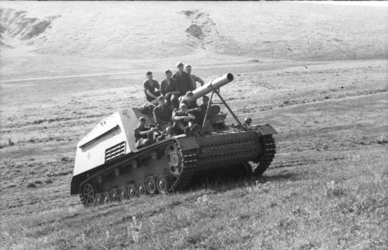 Black-and-white wartime photograph of a German Hummel self-propelled howitzer moving across open terrain with soldiers riding on it, its large 150mm howitzer in an open-topped fighting compartment.